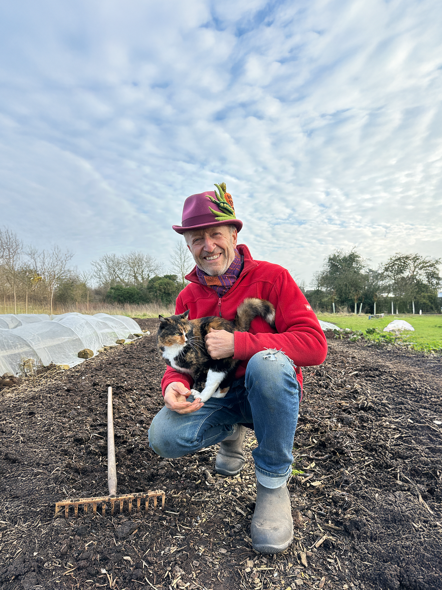 A man smiling, kneeling in a plant bed, holding a cat